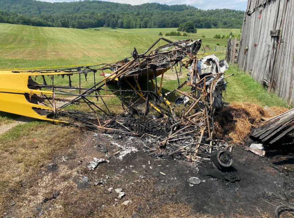 Close-up of the heavily charred and twisted remains of the Piper J3 Cub’s fuselage after crashing into a barn. Burn marks and scorched earth surround the destroyed aircraft frame.
